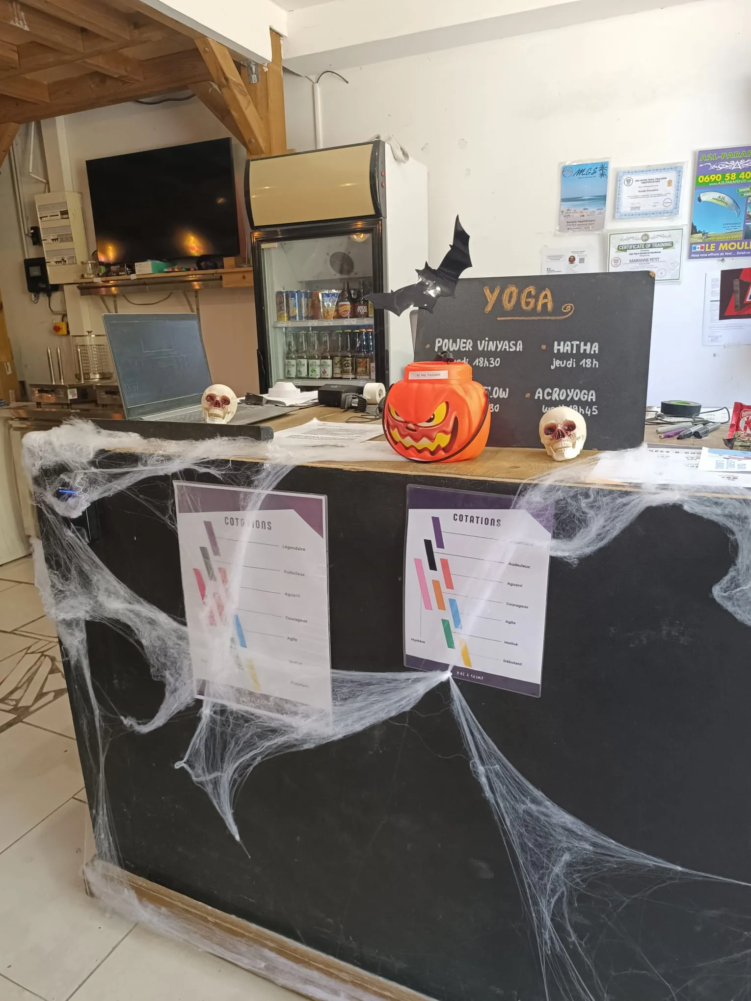 Comptoir d’accueil décoré pour Halloween : toiles d’araignée, citrouille grimaçante, crânes et chauve-souris devant le frigo à boissons